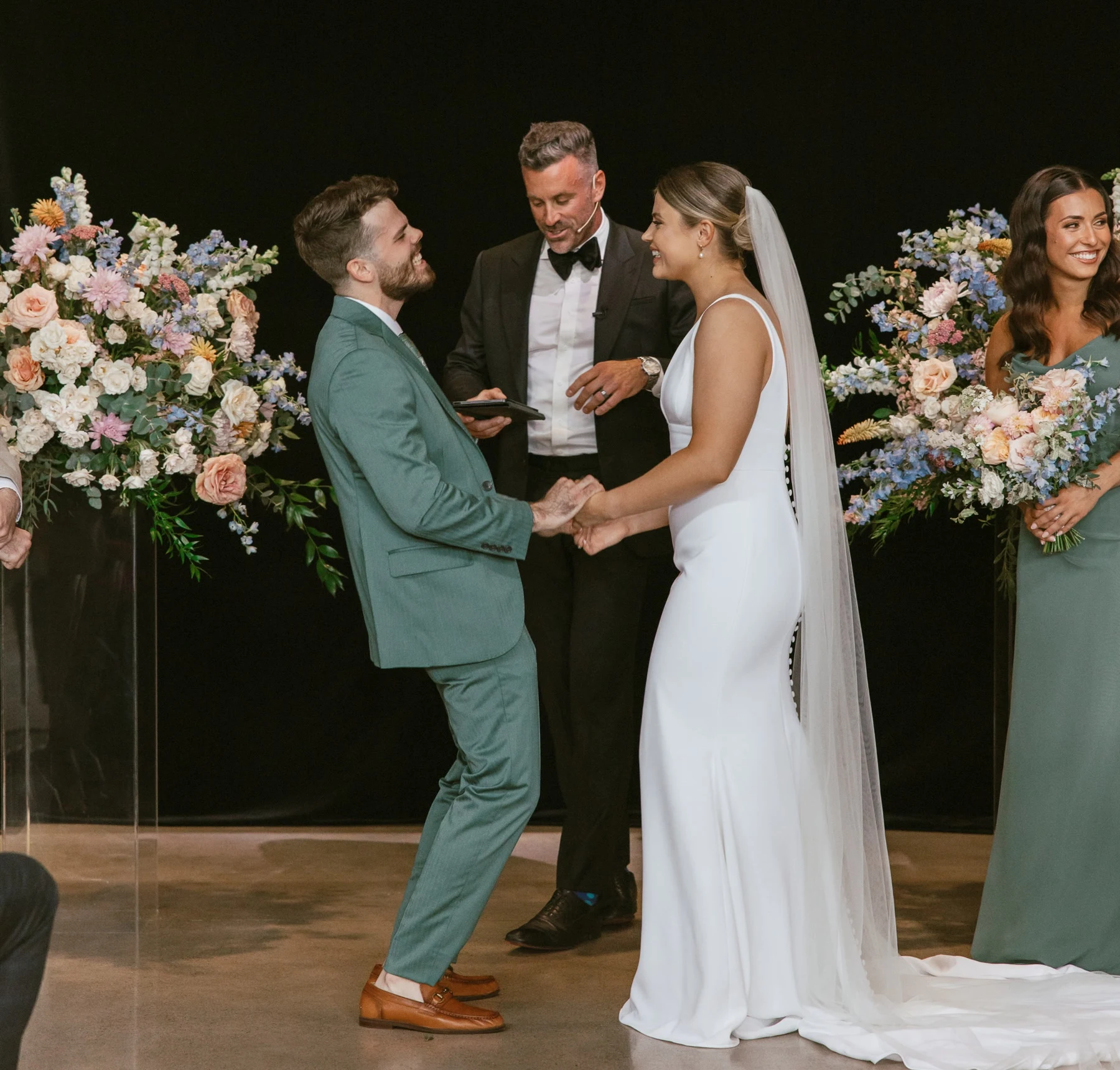 Jeff Maeck officiating a wedding ceremony as the smiling couple holds hands between colourful floral arrangements.